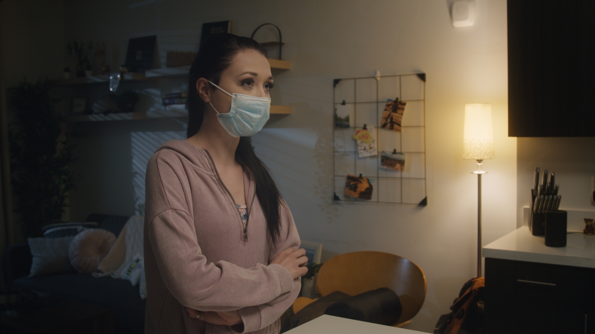 A young woman stands at her kitchen island. She is wearing a pink hoodie and a blue surgical mask. Her arms are folded defensively.