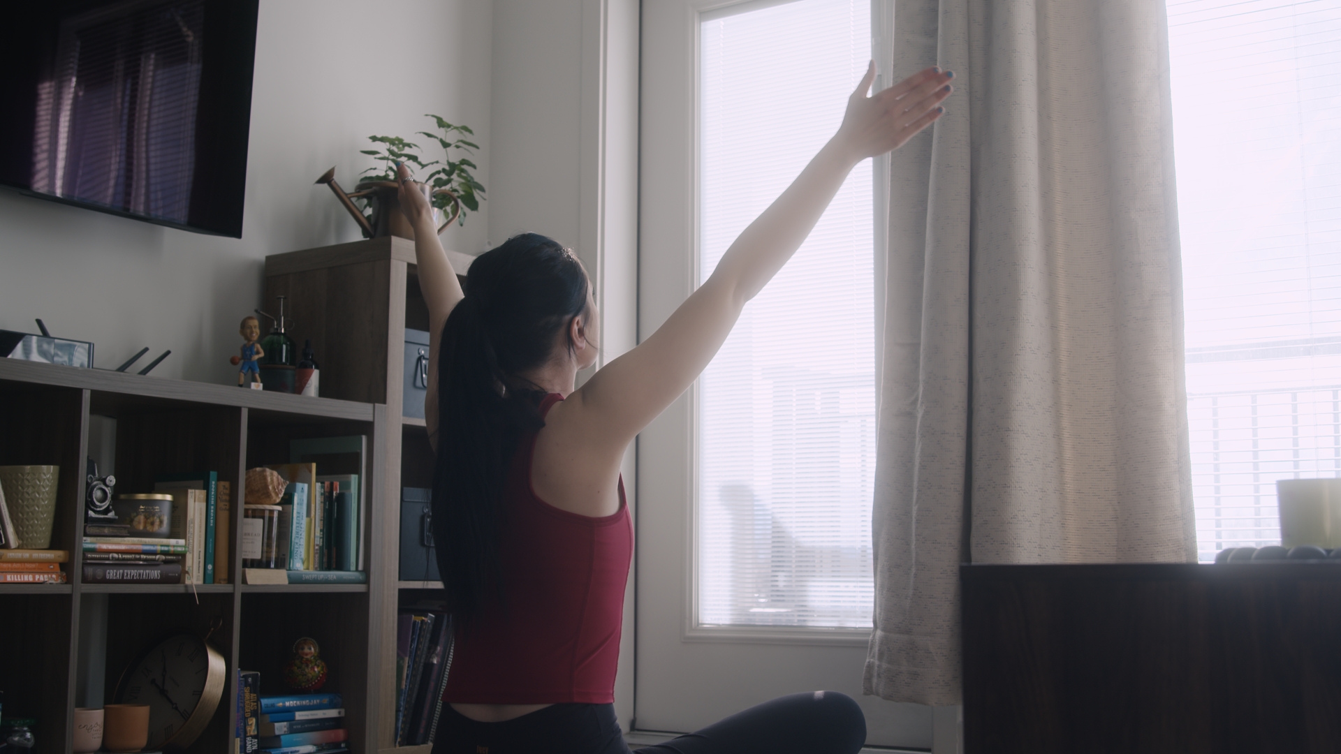 A young woman with long dark hair and a red tank top sits on the floor and raises her arms in a yoga pose in front of a bright window.