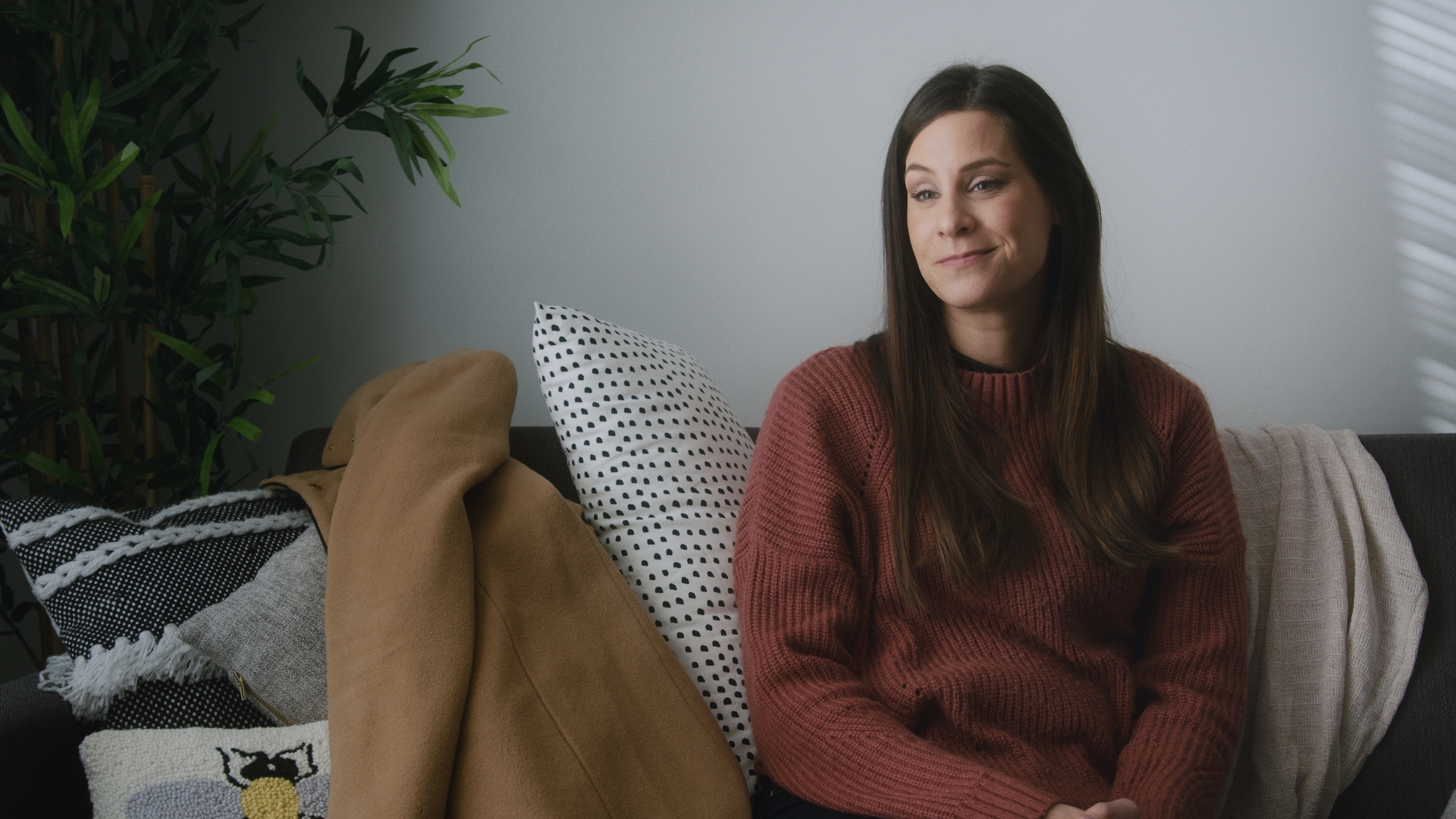 A woman with brown hair and a saffron sweater sits on a couch. She is smirking.