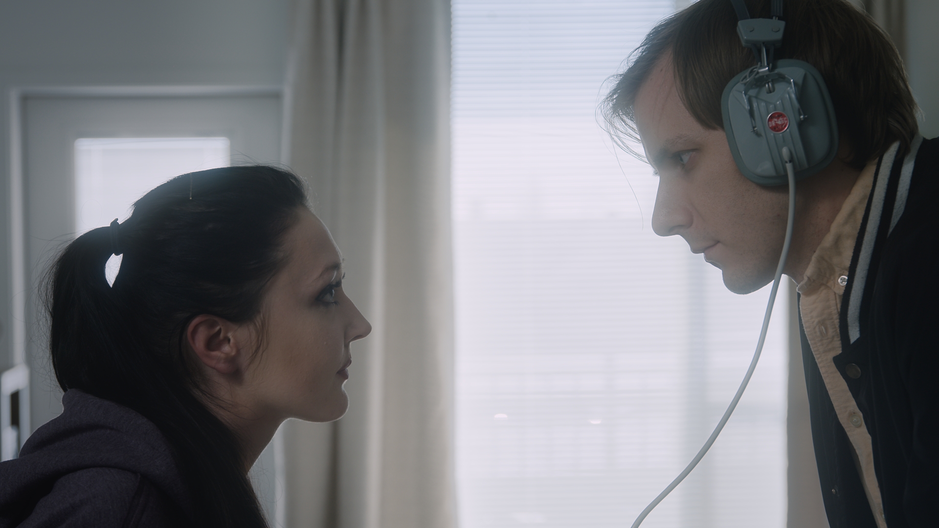 A woman with dark hair in a ponytail looks into the eyes of a young man with blond hair. The man is wearing old-fashioned headphones c. 1980s. The woman looks expectant as he listens.